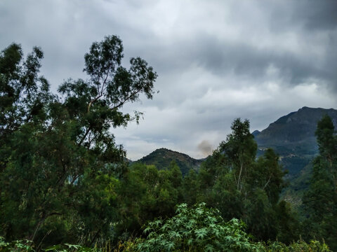 Lush green mountain ridges frame a distant puff of white smoke rising beneath a heavy overcast sky in this dramatic wide landscape photograph of nature