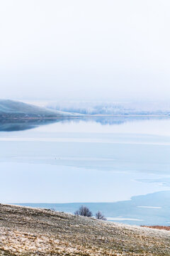Frosted trees and grass on the shore of lake at foggy sunrise. Trees and hills reflected in the water. Ice on the lake.