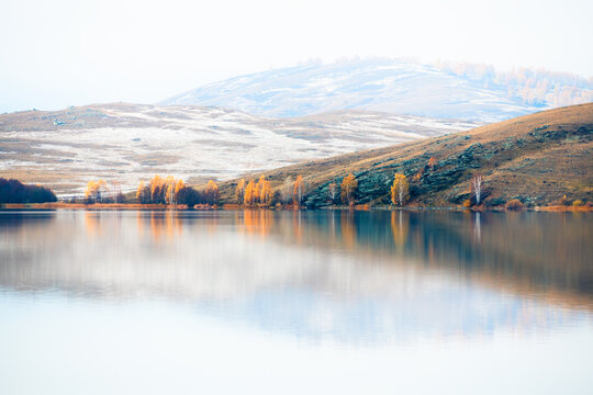 Snow-covered mountains with yellow autumn trees are reflected in the lake at foggy sunrise. Bashkortostan, Russia