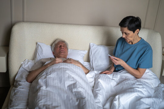 High angle view of worried older woman checking fever of sick husband in bed using thermometer. Illness management, marital responsibility, domestic healthcare, health awareness, supportive couple