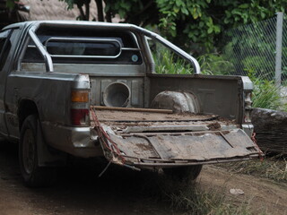 rear angle shot of an old, heavily used silver pickup truck parked on a dirt trail in a rural environment. The open tailgate, dirty truck bed, and worn body highlight themes of hard work, rural transp © นิรมล บุญเทพ