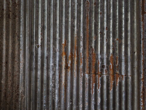 full-frame photograph of a corrugated galvanized iron sheet showing vertical ridges, silver metallic luster, and heavy orange-brown rust streaks. The image conveys themes of industrial aging, weathere