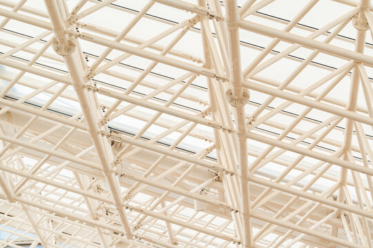 Vertical low angle view of a modern white steel space truss roof structure with translucent glass ceiling panels allowing soft natural light, highlighting geometric engineering design.