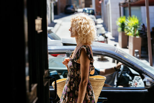 Blonde Woman Walking with Straw Bag Past Parked Car on Sunny Street During Summer in Spain
