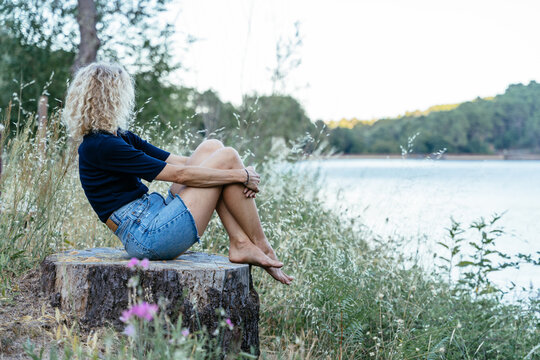 Mature Blonde Woman Sitting Barefoot by Peaceful Lake Enjoying Quiet Summer Moment in Nature