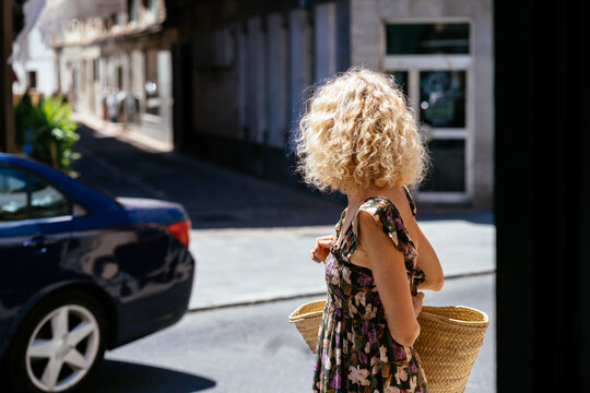 Blonde Woman Carrying Straw Bag While Walking Through Sunny Street in Small Spanish Town