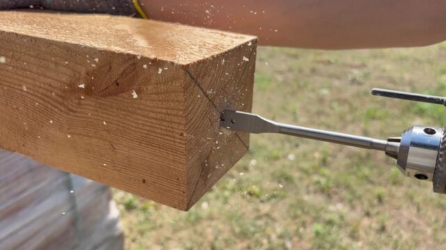 Close-up of a power drill with a spade bit drilling a hole into a thick wooden beam outdoors.