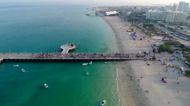 High angle aerial view shows lively Kish Island beach with clear turquoise water pier and people enjoying tropical seaside under sunny sky.