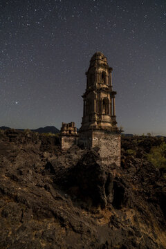 View of the historic bell tower of the San Juan Parangaricutiro church rising above the volcanic lava field under a clear starry night sky in Angahuan, Michoacan, Mexico.