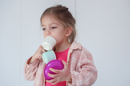 Portrait Of Young Girl Holding Colorful Stacking Cups And Touching One Cup With Mouth, Wearing Pink Plaid Shirt Against Bright Minimal Background, Showing Quiet Concentration, Playtime