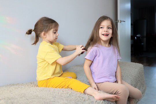 Two young girls playing indoors on soft shaggy rug, one carefully braiding other girl hair while smiling child sits nearby in bright home room with simple daylight and gentle family mood