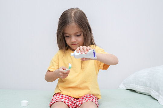 Young Girl Carefully Squeezes Toothpaste Onto Toothbrush While Sitting Indoors Against Clean White Background, Showing Focused Expression, Casual Clothing, Soft Daylight