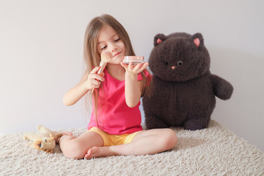 Young Girl Sits On Fluffy Rug Applying Makeup With Powder Brush And Compact Beside Large Plush Teddy Bear, Showing Playful Pretend Beauty Routine In Bright Minimal Bedroom With Soft Light
