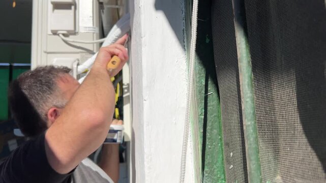 Horizontal video showing a male technician using a putty knife to apply sealant and cover the hole in a white exterior wall around the insulated pipes of a newly installed air conditioning unit.