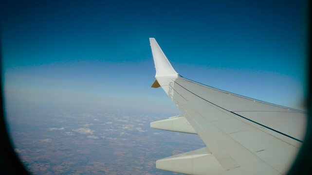 View through airplane window above scattered cumulus clouds and distant landscape. Aircraft wing against deep blue sky at cruising altitude. Commercial flight and aerial travel perspective