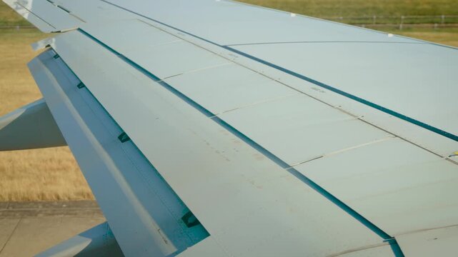 Runway taxiing with airplane wing, extended spoilers and flaps beside dry airport grass. Passenger window perspective before departure. Ground operations and commercial aviation
