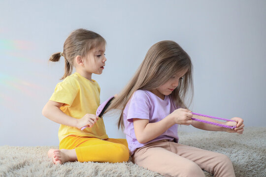 Two young girls sitting on soft carpet, one brushing long hair of friend while other holds purple braid accessory, calm indoor childhood grooming moment with pastel clothing and bright natural light