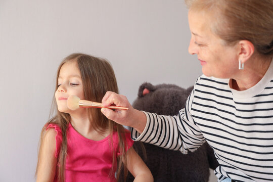 Close View Of Older Woman Using Makeup Brush On Childs Cheek Indoors, Showing Tender Grooming, Calm Expression, Casual Clothing, Soft Daylight, Simple Background, Selective Focus