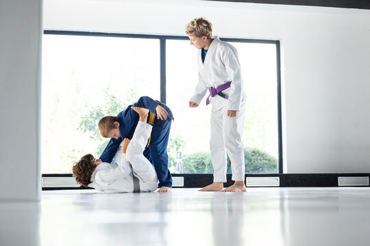 Female judo instructor supervises two young students practicing controlled ground grappling on dojo mat. Concept of safe sparring, technique correction and youth sports education.