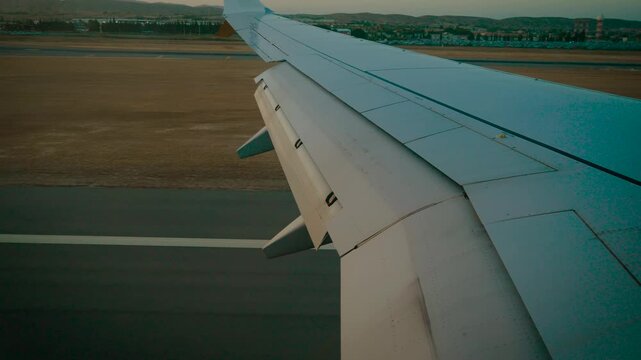 Touchdown approach with airplane wing and fully extended landing flaps over airport grounds. Passenger window view seconds before landing. Commercial jet descending toward runway in evening light