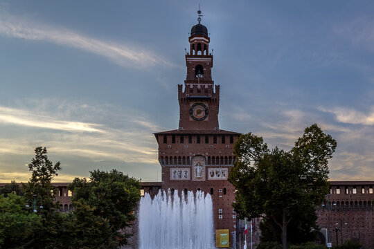 View of Sforzesco Castle with its iconic brick tower and clock, featuring a fountain in the foreground under a soft evening sky in Milano, Lombardia, Italy.