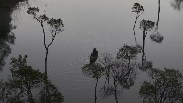 Aerial drone view of a fisherman rowing across Tuyen Lam Lake at sunrise near Suoi Tia in Xuan Huong, Da Lat, Vietnam, surrounded by peaceful pine forests.