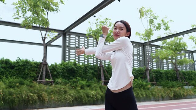 Young Asian sportswoman doing arm stretching exercise on red running track in outdoor park. Workout preparation, cool down stretch, fitness training, and wellness concept.