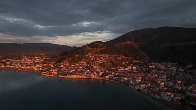 Drone aerial shot pulling back over the sea to reveal the historic coastal town of Nafpaktos with its Venetian port, castle hill, and golden sunrise light beneath dramatic sky in Greece.