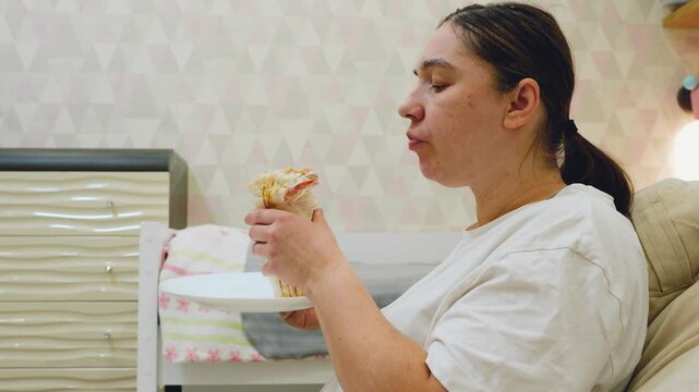 Adult woman in white shirt eating dessert from plate beside bed in warm home bedroom, shown in side profile with relaxed seated posture, soft fabrics, patterned wall and calm domestic mood