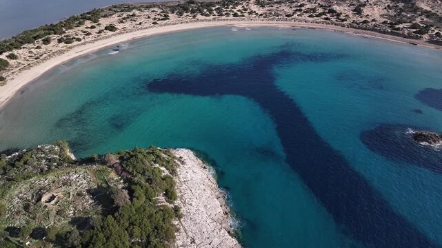 High-angle drone shot looking down at the vibrant turquoise waters, sandy curve, and rocky promontory of Voidokilia Beach in Messenia, Peloponnese, Greece, on a bright sunny day.