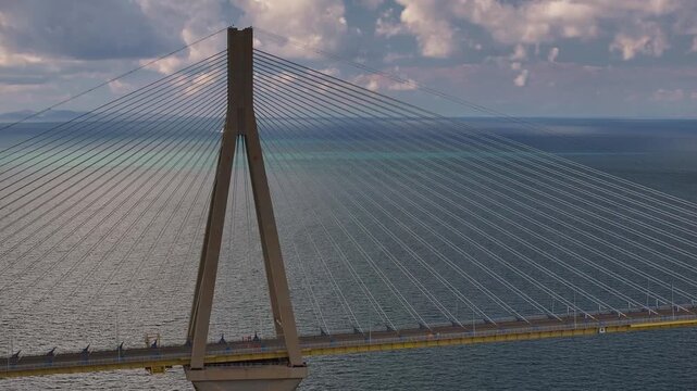 High-angle aerial shot focusing on the main pylon and stay cables of Rio-Antirrio bridge, as a red semi-trailer truck drives across the deck over the sea on a sunny cloudy day in Greece.
