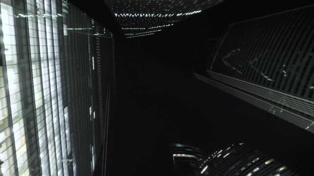 Upward rotating view looks between reflective Moscow skyscrapers where bright office windows form diagonal grids against black sky and the close glass architecture creates a dramatic urban canyon