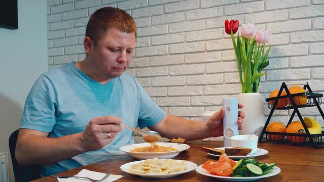 Adult man eating home meal at wooden dining table with pasta, vegetables, bread, fruit, tulips, and white brick wall in calm indoor domestic setting, with man, adult, eating, meal detail