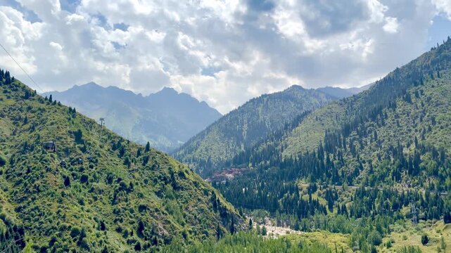 4K panoramic view of lush green mountains near Almaty, Kazakhstan, seen from Kok Tobe viewpoint. Scenic mountain valley, forested slopes and dramatic cloudy sky create a beautiful summer landscape.