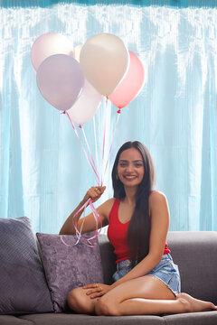 Young woman holding bunch of balloons while sitting on  sofa in living room