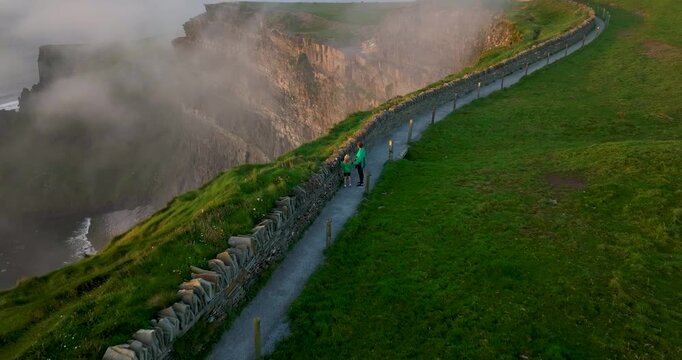 Aerial drone view of mother and daughter walking along Cliffs of Moher coastal path at sunset, Ireland