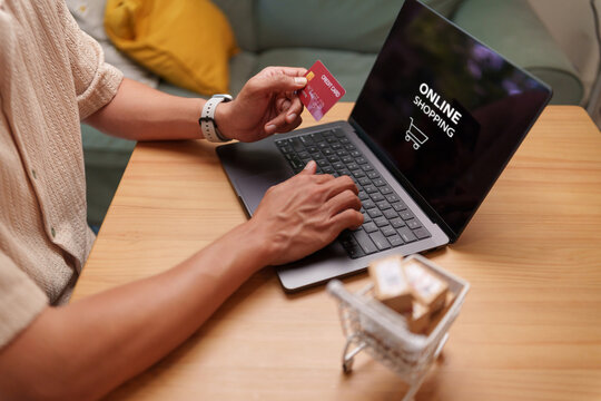 Man holding a credit card while shopping online on laptop with a mini cart and packages, representing e-commerce checkout, digital payment, retail purchasing and convenient home delivery.