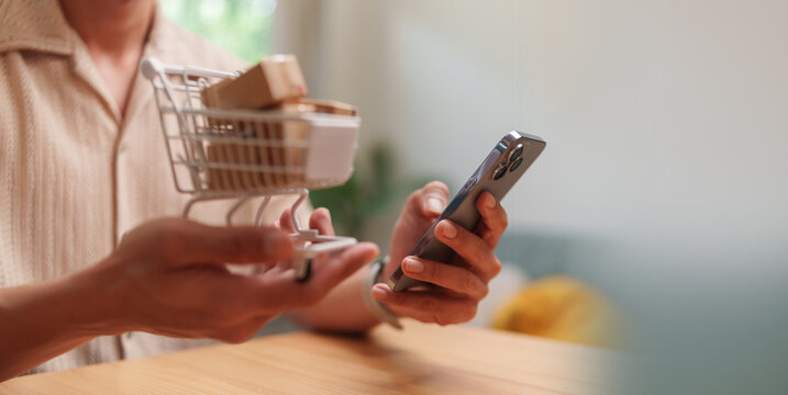 Man holding a mini shopping cart with packages while using a smartphone for online shopping, representing mobile e-commerce, digital purchasing, delivery