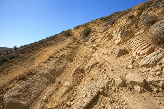 Steep unpaved trail on dry rocky hillside with ruts and exposed roots, arduous hiking path concept