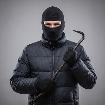 A criminal wearing a black balaclava and holding a metal crowbar against a gray background.