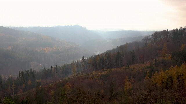 Panoramic autumn view over Karlovy Vary valley in Czechia, with forested hills fading into soft morning haze. Scenic spa region landscape shows colorful woodland, slopes, mist and distant ridges
