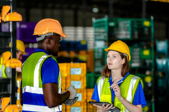 Black male logistics employee and his female coworker wearing hard hats and hi-vis vests verify bin locations across the parts supermarket at an automotive parts production plant with.