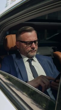 Vertical video of middle aged Caucasian businessman sitting in back seat of luxury car and using smartphone. Clip is ideal for corporate travel, executive lifestyle, and mobile business solutions