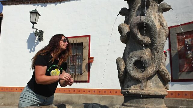 Woman tourist refreshing herself with flowing water from Pila de las Culebras serpent fountain in Tapalpa Pueblo M&aacute;gico, Jalisco, Mexico. Travel lifestyle shot.