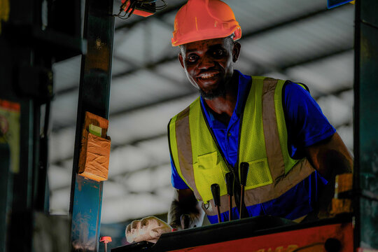 Black male logistics employee wearing an orange protective helmet and hi-vis clothing works the hydraulic controls of warehouse machinery in a large fulfilment centre with rows of stacked p.