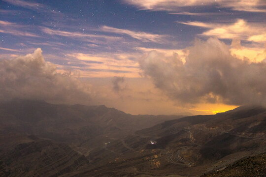 View of Jebel Jais mountain landscape at night with a winding road, starry sky, and clouds illuminated by a warm horizon glow in Ras Al Khaimah, United Arab Emirates.
