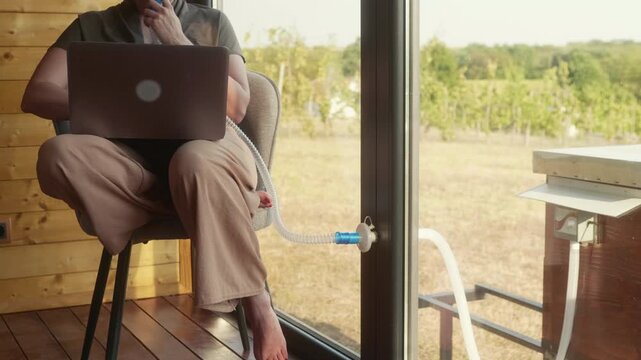 Focused middle-aged woman works on a laptop while breathing through a mask during an apitherapy session inside a beehive air chamber.