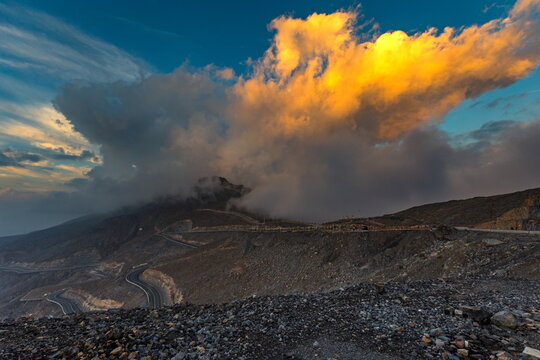 View of the winding mountain road at Jebel Jais, featuring dramatic golden clouds over the rugged rocky landscape of Ras Al Khaimah, United Arab Emirates.
