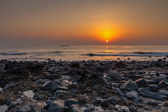 View of a rocky beach at sunset with calm waves gently lapping against dark stones under a vibrant orange sky in Ras Al-Khaimah, Ras Al Khaimah, United Arab Emirates.
