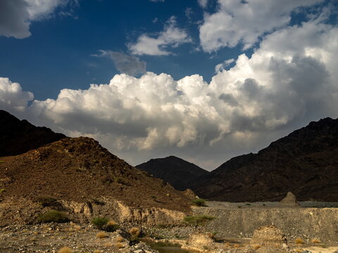 View of the rugged Hajar Mountains landscape with rocky terrain, sparse desert vegetation, and dramatic white clouds against a deep blue sky in Ras Al-Khaimah, Ras Al Khaimah, United Arab Emirates.